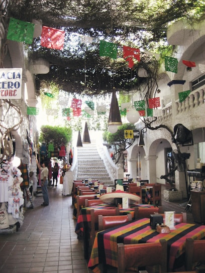 A charming restaurant interior with colorful tablecloths and rustic wooden chairs arranged in a dining area. Brightly colored papel picado flags hang overhead, adding a festive touch. The ceiling is adorned with greenery, giving a natural and inviting ambiance. A white staircase leads to an upper level, flanked by decorative white stucco walls and arches. Various clothing items hang on display to the left, and people are casually browsing.