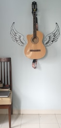 A wooden acoustic guitar is mounted against a light blue wall decorated with stylized black and white wing decals on either side of the guitar. There is a dark wooden chair with a light cushioned seat positioned to the left, which holds a closed gray laptop.