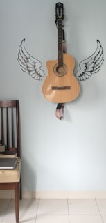 A wooden acoustic guitar is mounted against a light blue wall decorated with stylized black and white wing decals on either side of the guitar. There is a dark wooden chair with a light cushioned seat positioned to the left, which holds a closed gray laptop.