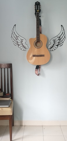 A wooden acoustic guitar is mounted against a light blue wall decorated with stylized black and white wing decals on either side of the guitar. There is a dark wooden chair with a light cushioned seat positioned to the left, which holds a closed gray laptop.