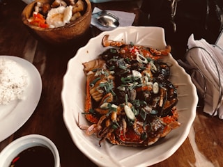 A beautifully arranged seafood dish featuring crabs, garnished with green onions and a colorful sauce, served on a large white platter. Nearby, a bowl of rice and a wooden bowl containing various foods, including a fried egg and diced tomatoes, are present. On the table, a cup with a dark liquid and a set of napkins complete the setting.