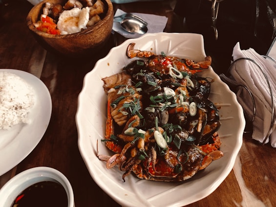 A beautifully arranged seafood dish featuring crabs, garnished with green onions and a colorful sauce, served on a large white platter. Nearby, a bowl of rice and a wooden bowl containing various foods, including a fried egg and diced tomatoes, are present. On the table, a cup with a dark liquid and a set of napkins complete the setting.