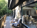 A wooden deck extends alongside a house with large glass windows. There are several wooden chairs and a small table with potted plants on the deck. Inside the house, a person is visible working on a laptop. The surrounding area is filled with dense green trees and sunlight filters through, creating a serene atmosphere.
