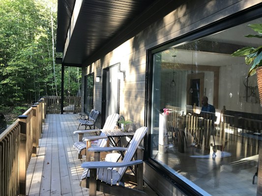 A wooden deck extends alongside a house with large glass windows. There are several wooden chairs and a small table with potted plants on the deck. Inside the house, a person is visible working on a laptop. The surrounding area is filled with dense green trees and sunlight filters through, creating a serene atmosphere.