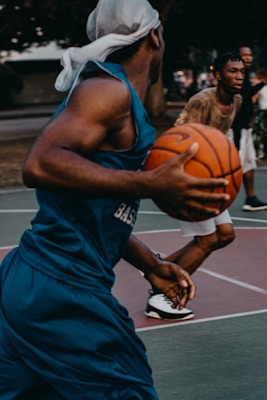 Two people are playing basketball outdoors on a court. One player is in the foreground wearing a blue jersey and holding a basketball, while the other player in the background is wearing casual clothing. The scene captures dynamic movement and action, showing the intensity of a street basketball game.