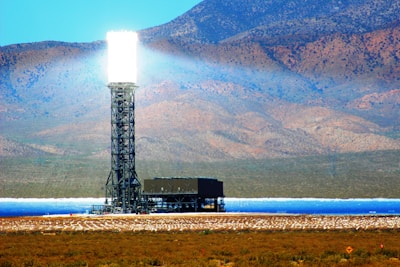 A large heliostat tower is situated in a desert landscape with mountains in the background. The tower emits a very bright, intense light at the top which likely comes from concentrated solar power. Below the tower is what appears to be part of a solar power facility including numerous mirrors that reflect sunlight to the top of the tower. The surrounding terrain is arid and rocky, with sparse vegetation.