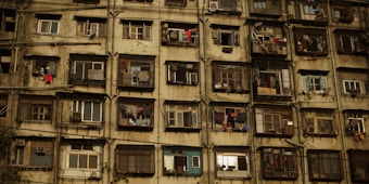 A large, weathered apartment building facade with multiple rectangular windows, some of which have open shutters or clothes hanging out to dry. The building appears old and worn, with visible signs of aging and disrepair.