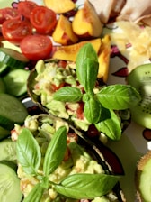 A vibrant arrangement of fresh foods including halved avocados filled with guacamole, topped with fresh basil leaves. Surrounding them are slices of cucumber, cherry tomatoes, peach slices, kiwi, and some sliced cheese or chicken.
