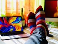 A pair of feet in colorful, striped socks is resting on a desk. The desk features an open laptop displaying a vibrant, abstract image. Light streams through a window with sheer curtains, casting shadows. There is also a blurred monitor and a bottle in the background.