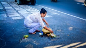 A person in traditional attire is placing an offering on the ground by the side of a road. The offerings include flowers, food, and incense sticks arranged on a small platform. The street is paved, and a few vehicles can be seen in the background.