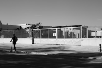 A black and white image features an industrial facility with large metal gates labeled 'Puerta 1'. A person on a scooter rides along the sidewalk in the foreground, casting a shadow on the ground. The industrial building and equipment can be seen in the background, enclosed by a high fence.