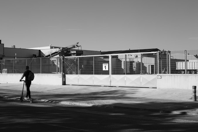 A black and white image features an industrial facility with large metal gates labeled 'Puerta 1'. A person on a scooter rides along the sidewalk in the foreground, casting a shadow on the ground. The industrial building and equipment can be seen in the background, enclosed by a high fence.