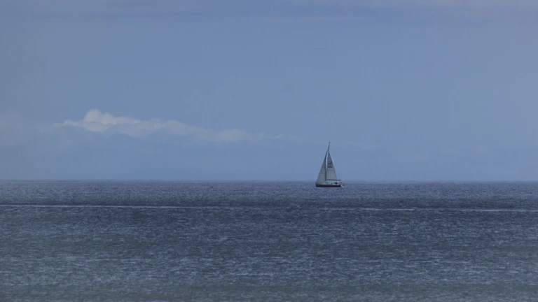 Calm waters reflecting a cloudy sky with a lone sailboat drifting