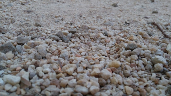 A close-up view of a gravel surface composed of small, irregularly shaped stones and pebbles in varying shades of beige, gray, and brown. The image captures the texture and unevenness of the surface with some focus on the foreground.