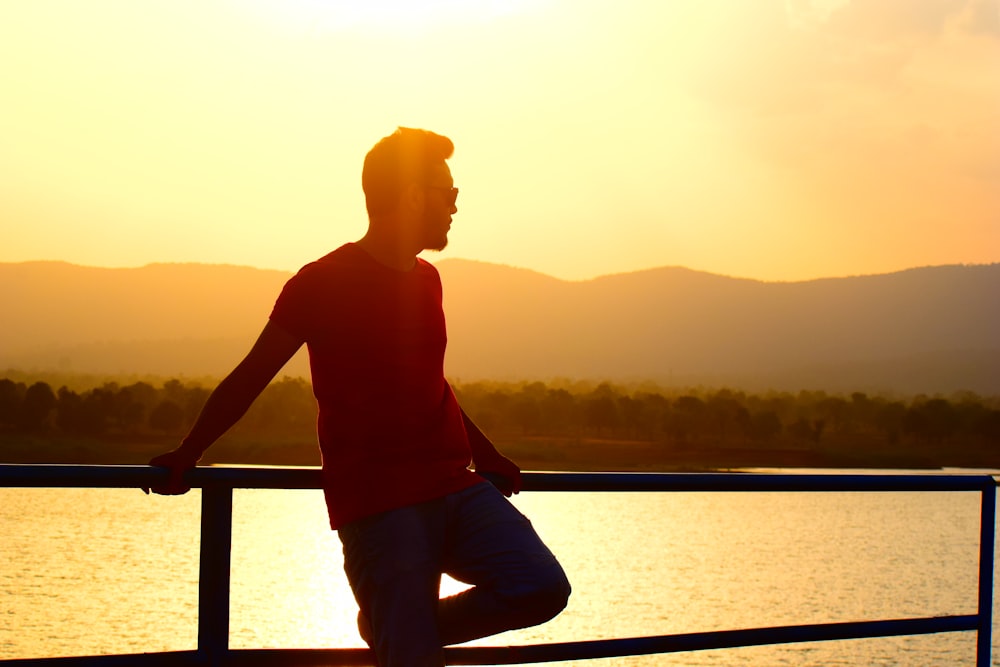 man in red t-shirt and black pants leaning on railing in beach at sunset