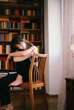 A person with long hair sits on a wooden chair, arms wrapped around their knees, in a cozy room with a background of colorful bookshelves. Soft light filters through a curtain, creating a calm and introspective atmosphere.