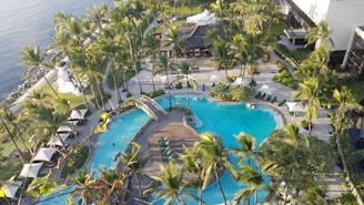 An aerial view captures a luxurious resort area featuring a large swimming pool surrounded by palm trees. The pool has a unique shape with a central island connected by small bridges. Sun loungers with umbrellas line the poolside. To the left, a lawn overlooks a body of water, with a lighthouse structure visible. The entire setting exudes a tropical and serene atmosphere.