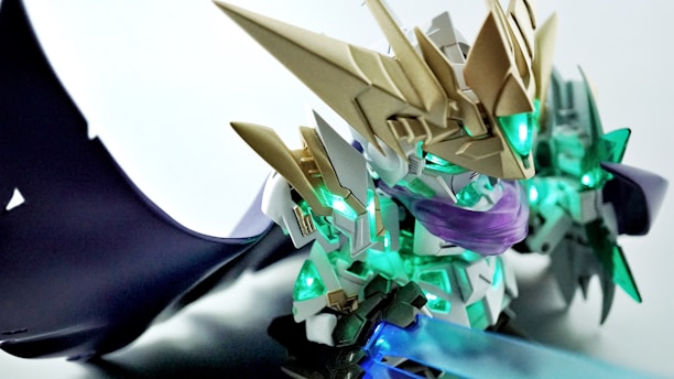 A highly detailed model figure of a robot or armored character, featuring intricate golden armor, glowing green accents, and a translucent weapon. The robot has a dynamic pose, with elaborate wings or extensions in the background, giving a sense of motion.