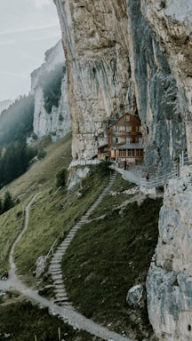 A rustic wooden house is built into the side of a towering cliff, surrounded by lush greenery and rugged rock formations. The house is accessible by a narrow path and steps cut into the hillside. Misty hills and a forested landscape stretch into the background, creating a serene and isolated atmosphere.