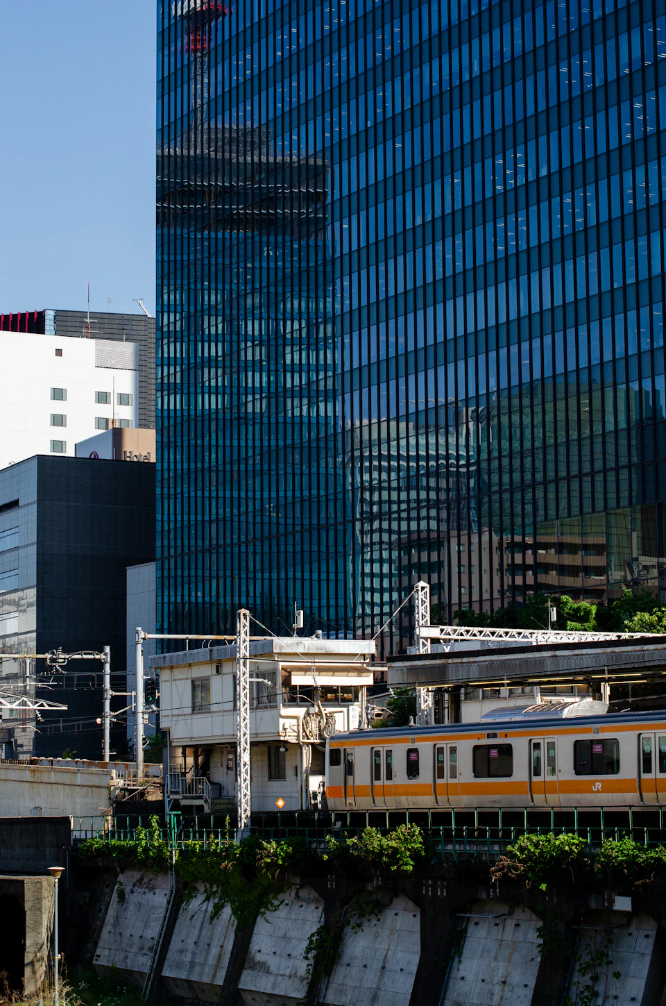 The image depicts a bustling city scene with a train station and tall buildings in the background, including a large blue glass skyscraper that reflects the clear sky above. A white train is visible on the tracks, adding to the urban atmosphere. The perspective of the photo is taken from across the street, providing a comprehensive view of the cityscape.
