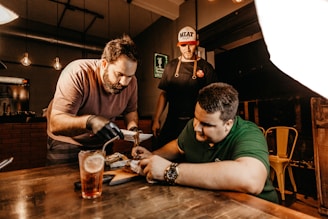 Three men are gathered around a wooden table in a warm, dimly lit setting. One man, wearing a pink shirt and gloves, is focused on serving food. Another man, in a green shirt, appears to be closely inspecting or interacting with the dish. A third man stands in the background, wearing a cap and apron, observing the scene. A glass of iced drink with a lemon slice sits on the table, adding to the casual dining atmosphere.
