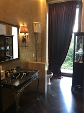 A luxurious bathroom with ornate decor, including a black marble sink with gold fixtures and a decorative mirror. The walls are adorned with a patterned wallpaper, and a vintage-style wall lamp adds a touch of elegance. A high-level cistern toilet is set next to a window dressed with heavy, dark curtains. To the side, a cabinet displays several small items.