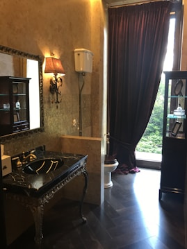 A luxurious bathroom with ornate decor, including a black marble sink with gold fixtures and a decorative mirror. The walls are adorned with a patterned wallpaper, and a vintage-style wall lamp adds a touch of elegance. A high-level cistern toilet is set next to a window dressed with heavy, dark curtains. To the side, a cabinet displays several small items.