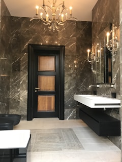 A modern bathroom with dark marble walls and a black wooden door with lighter wood panels. A chandelier and several wall sconces provide warm lighting, reflected in mirrors mounted above a sleek, wall-mounted white sink. The floor is tiled in light gray, contrasting with the dark tones of the room.