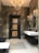 A modern bathroom with dark marble walls and a black wooden door with lighter wood panels. A chandelier and several wall sconces provide warm lighting, reflected in mirrors mounted above a sleek, wall-mounted white sink. The floor is tiled in light gray, contrasting with the dark tones of the room.