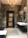A modern bathroom with dark marble walls and a black wooden door with lighter wood panels. A chandelier and several wall sconces provide warm lighting, reflected in mirrors mounted above a sleek, wall-mounted white sink. The floor is tiled in light gray, contrasting with the dark tones of the room.