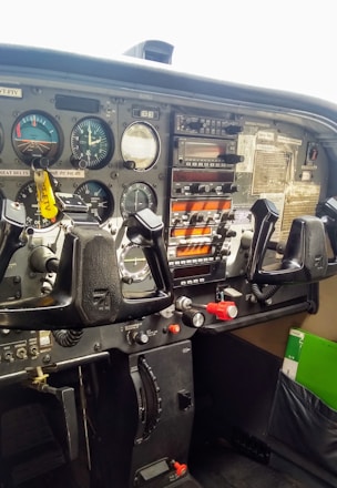 An airplane cockpit featuring various flight instruments and controls. The dashboard contains multiple dials, gauges, and switches, with a prominent control yoke in front. The colors on the instruments display important flight data. There is a checklist and other documents clipped to the right side.