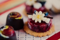 A close-up view of an assortment of artisanal desserts, including a cookie topped with a glossy berry glaze and an edible flower, alongside colorful, marbled chocolates.