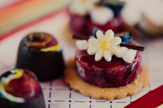 A close-up view of an assortment of artisanal desserts, including a cookie topped with a glossy berry glaze and an edible flower, alongside colorful, marbled chocolates.