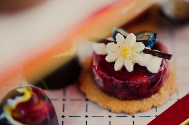 A beautifully presented dessert featuring a small round cake topped with a glossy red glaze. Decorated with a white flower, blueberry pieces, and thin chocolate sticks, all placed on a biscuit base. Nearby, a dark chocolate confection showcases a swirl of vibrant colors.