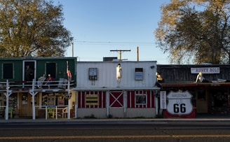 A row of vintage-style storefronts, likely part of a Route 66 attraction, featuring mannequins dressed in retro clothing and a large Route 66 sign. The area is adorned with quaint wooden structures with signs for shops and souvenirs. The scene is framed by large trees, and the light suggests late afternoon or early evening.