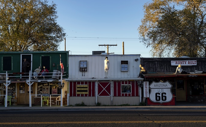 A row of vintage-style storefronts, likely part of a Route 66 attraction, featuring mannequins dressed in retro clothing and a large Route 66 sign. The area is adorned with quaint wooden structures with signs for shops and souvenirs. The scene is framed by large trees, and the light suggests late afternoon or early evening.
