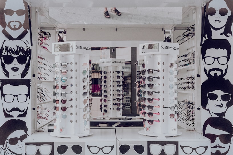 A sunglasses store display with multiple stands showcasing various styles of sunglasses. The walls are adorned with stylized black and white illustrations of people wearing sunglasses. The stands are cylindrical and display both aviator and sporty designs in different colors. The surrounding area appears to be well-lit, reflecting a modern retail environment.
