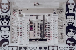 A sunglasses store display with multiple stands showcasing various styles of sunglasses. The walls are adorned with stylized black and white illustrations of people wearing sunglasses. The stands are cylindrical and display both aviator and sporty designs in different colors. The surrounding area appears to be well-lit, reflecting a modern retail environment.