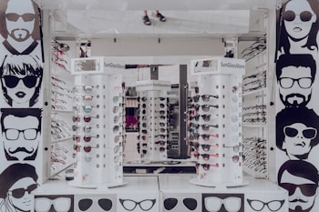 A sunglasses store display with multiple stands showcasing various styles of sunglasses. The walls are adorned with stylized black and white illustrations of people wearing sunglasses. The stands are cylindrical and display both aviator and sporty designs in different colors. The surrounding area appears to be well-lit, reflecting a modern retail environment.