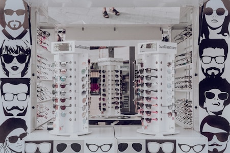 A sunglasses store display with multiple stands showcasing various styles of sunglasses. The walls are adorned with stylized black and white illustrations of people wearing sunglasses. The stands are cylindrical and display both aviator and sporty designs in different colors. The surrounding area appears to be well-lit, reflecting a modern retail environment.