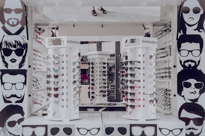 A sunglasses store display with multiple stands showcasing various styles of sunglasses. The walls are adorned with stylized black and white illustrations of people wearing sunglasses. The stands are cylindrical and display both aviator and sporty designs in different colors. The surrounding area appears to be well-lit, reflecting a modern retail environment.