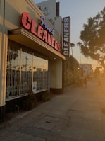 A dry cleaning shop with a prominent red and blue neon sign saying 'French CLEANERS' is situated along a pedestrian sidewalk. The building has large glass windows with metal bars. On the side, there are some potted plants. The scene is bathed in warm, golden sunlight, indicative of either sunrise or sunset. There are a few people walking down the street, casting long shadows.