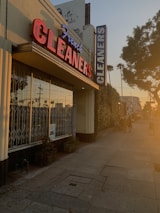 A dry cleaning shop with a prominent red and blue neon sign saying 'French CLEANERS' is situated along a pedestrian sidewalk. The building has large glass windows with metal bars. On the side, there are some potted plants. The scene is bathed in warm, golden sunlight, indicative of either sunrise or sunset. There are a few people walking down the street, casting long shadows.