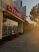 A dry cleaning shop with a prominent red and blue neon sign saying 'French CLEANERS' is situated along a pedestrian sidewalk. The building has large glass windows with metal bars. On the side, there are some potted plants. The scene is bathed in warm, golden sunlight, indicative of either sunrise or sunset. There are a few people walking down the street, casting long shadows.