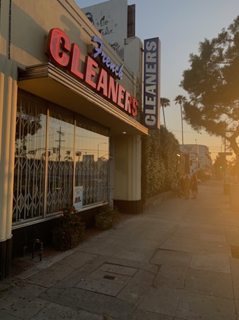 A dry cleaning shop with a prominent red and blue neon sign saying 'French CLEANERS' is situated along a pedestrian sidewalk. The building has large glass windows with metal bars. On the side, there are some potted plants. The scene is bathed in warm, golden sunlight, indicative of either sunrise or sunset. There are a few people walking down the street, casting long shadows.
