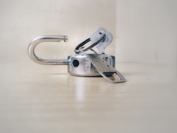 A metallic padlock is unlocked with keys inserted into it. The lock is lying on a light-colored surface with a blurred background. The keys are attached to a small metal key ring.