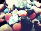 A collection of colorful stones with a variety of textures and shades, including pinks, blues, greens, and grays. The stones are irregularly shaped and placed closely together, creating an interesting and vibrant texture.