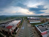 Aerial view of an urban shopping area with multiple retail and restaurant buildings. The setting appears calm, with an empty parking lot and a soft evening sky in the background. Buildings have red and beige tones with some greenery, and there's a prominent display of branded signs and logos.
