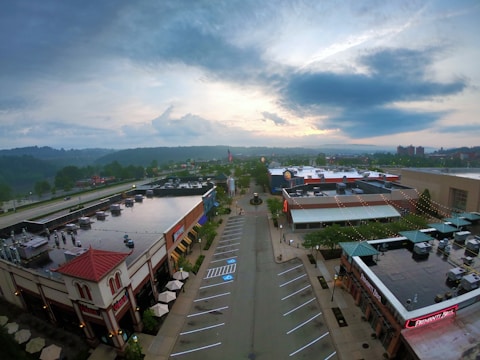 Aerial view of an urban shopping area with multiple retail and restaurant buildings. The setting appears calm, with an empty parking lot and a soft evening sky in the background. Buildings have red and beige tones with some greenery, and there's a prominent display of branded signs and logos.