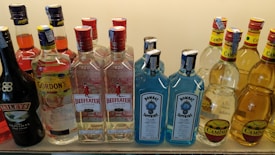 A collection of various liquor bottles including brands like Baileys, Aperol, Gordon's, Beefeater, Bombay Sapphire, and Camino. The bottles are arranged in rows on a shelf against a neutral background.